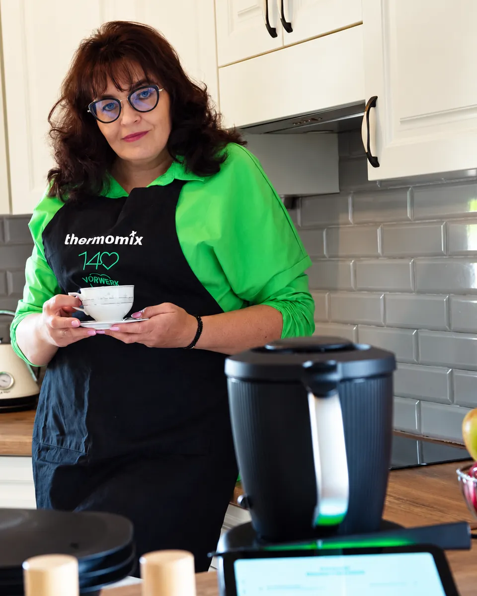 Bożena w fartuchu Thermomix z filiżanką w kuchni, na pierwszym planie urządzenie Thermomix