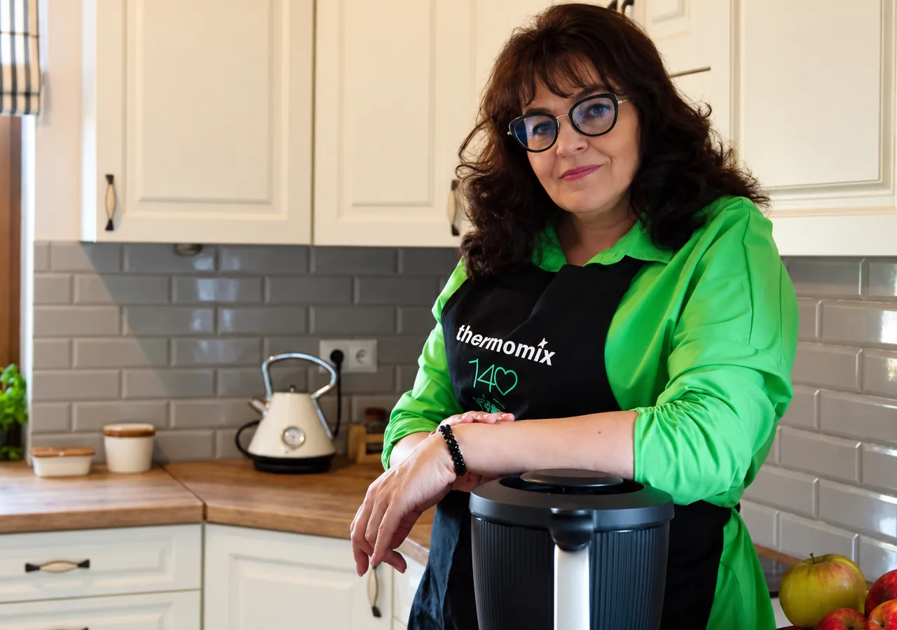 Bożena w zielonej koszuli i fartuchu Thermomix opiera się o urządzenie Thermomix w swojej kuchni